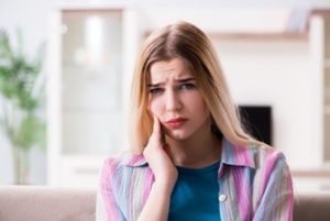 Woman sitting in a living room holding her cheek with a look a discomfort on her face.